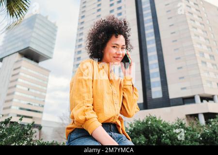 Young latin woman with curly hair having a call by smart phone. Hispanic lady with yellow shirt talking by cellphone sitting outdoors. Communication concept. High quality photo Stock Photo