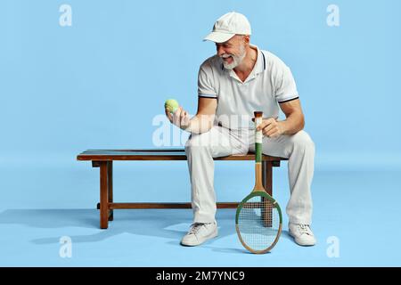 Portrait of handsome senior man in stylish white outfit sitting on bench with tennis ball and racket over blue background. Concept of leisure activity Stock Photo