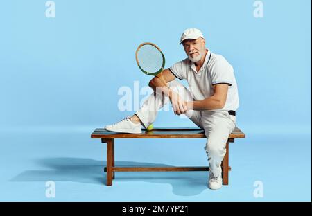 Popular game. Portrait of handsome senior man in stylish white outfit sitting on bench over blue background. Concept of leisure activity, hobby Stock Photo