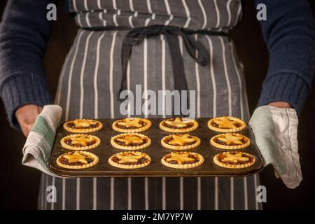 Closeup of the hands of a unidentifiable person wearing a traditional striped apron, holding a tray of hot, star topped minces pie, with a tea towel. Stock Photo