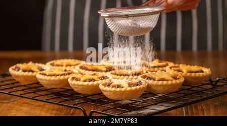 Mince pies topped with pastry stars on a metal rack being dredged in icing sugar by an unidentifiable person in a black striped apron. Stock Photo