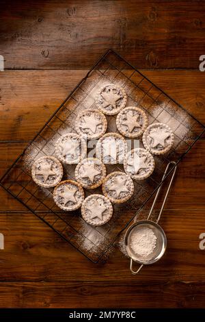 Flat lay mince pies topped with pastry stars, dusted with icing sugar on dark wooden background: Christmas home baking. Stock Photo