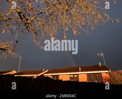 A feint rainbow in a dark grey sky over winter sunlit houses framed by leafless tree, Worcestershire, England. Stock Photo