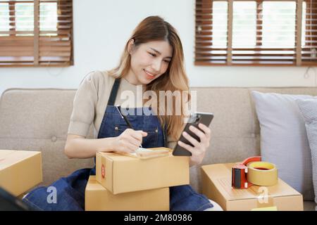 Starting small business entrepreneur of independent young Asian woman online seller is using smart phone and taking orders to pack products for Stock Photo