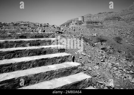 Black and white shot of stone walls of small medieval Arabian fort under tall mountain cliffs. Fortress in Bukha, Musandam peninsula, Oman. Hot, hazy Stock Photo