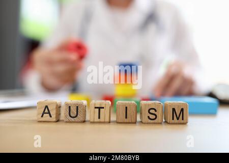 Word autism on wooden blocks close-up and blurred doctor with multi-colored blocks and toys in background. Stock Photo