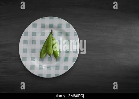 Three broad beans in shells on a round plate on a black wooden background with copy space. Stock Photo