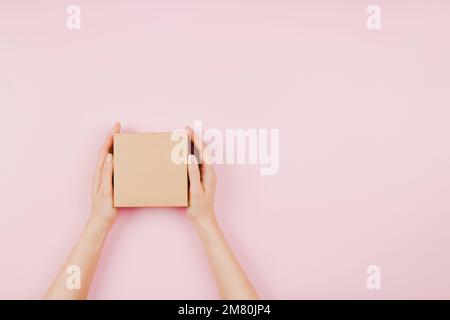 Top view to female hands holding small rectangle brown cardboard box on pastel pink background. Mockup parcel box. Packaging, shopping, delivery Stock Photo