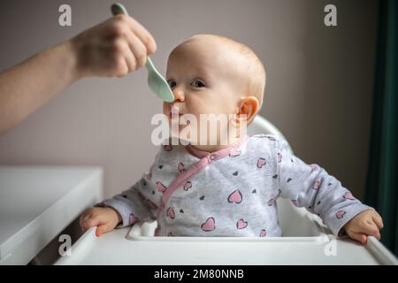 Baby girl eating blend mashed food sitting, on high chair, mother feeding child, hand with spoon for vegetable lunch, baby weaning, first solid food f Stock Photo