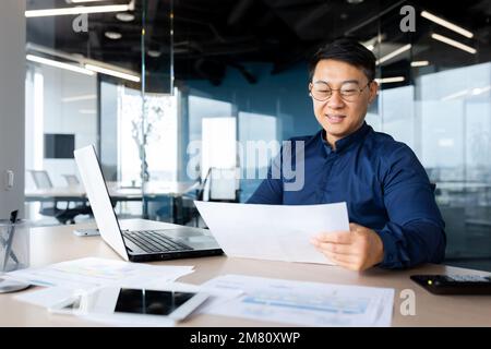 A young man, an Asian architect, designer, engineer, freelancer works in the office at a desk using a laptop. Holds documents, plans, drawings in his hands. Stock Photo