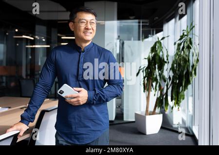 Portrait of a confident smiling Asian man. Businessman, director. He is standing in the office at the table, holding the chair with his hand, holding the phone in his hand, looking out the window. Stock Photo