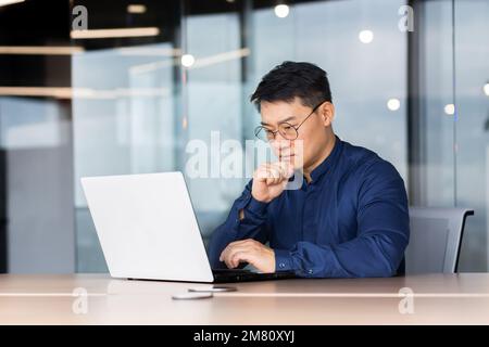 Young concentrated male Asian student studying at laptop. Sitting at the desk in the office, on campus, in the library. Takes an exam, online training via video call, listens to a lecture. Stock Photo