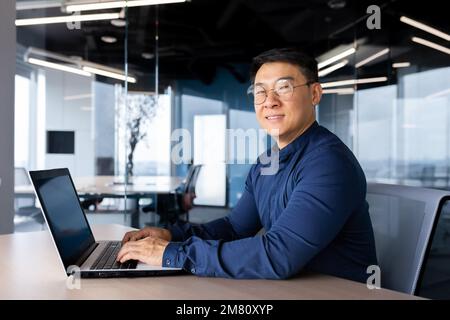 Portrait of a young male Asian student studying at a laptop. Sitting at the desk in the office, on campus, in the library. He studies online, remotely. He looks at the camera, smiles. Stock Photo