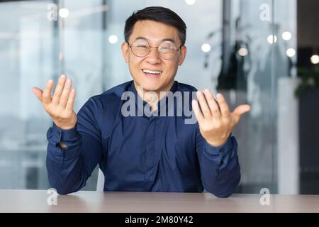 Portrait young male Asian teacher is lecturing students, taking exams online video call. Sitting in the office at the table, smiling at the camera, explains, waving his hands. Stock Photo
