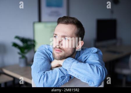 Cheerful bearded caucasian male sitting in home office looking at side dreaming. Young businessman or entrepreneur in denim shirt with computer monitor on background. High quality image Stock Photo