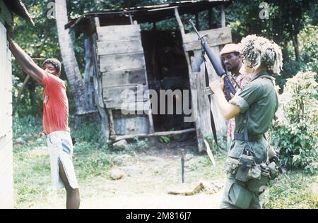 A member of the 82nd Airborne Division guards two prisoners as another soldier searches a small outbuilding during Operation URGENT FURY. Subject Operation/Series: URGENT FURY Country: Grenada (GRD) Stock Photo