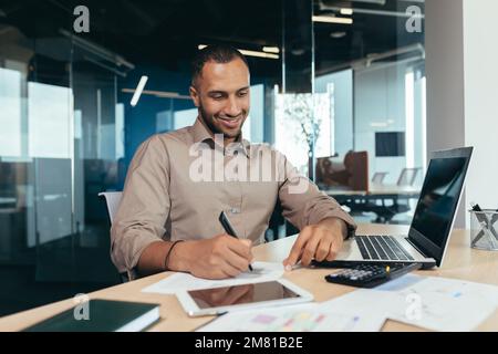 Successful african american businessman inside office doing paperwork, man writing information, worker using laptop for work sitting with bills and contracts smiling and satisfied with result. Stock Photo