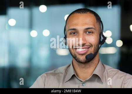 Close up hispanic male worker with video headset smiling and looking at camera, tech support worker working inside office. Stock Photo