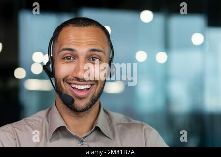 Close up hispanic male worker with video headset smiling and looking at camera, tech support worker working inside office. Stock Photo