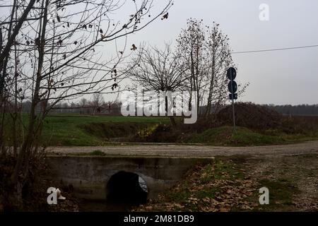 Crossing  between a dirt trail and a paved one next to a stream of water on a cloudy day in the italian countryside Stock Photo