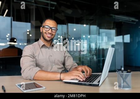 Portrait of successful hispanic businessman inside office, man with laptop working typing on keyboard smiling and looking at camera. Stock Photo