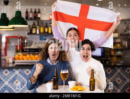 Company of young adult emotional fans supporting English team with state flag while resting in sports bar with beer Stock Photo