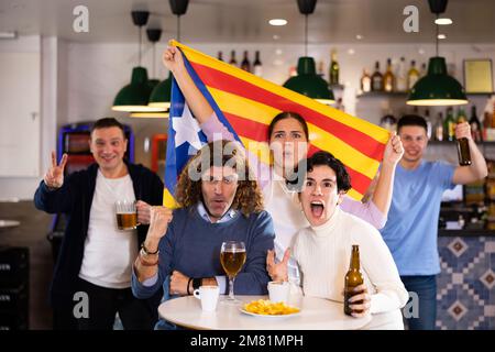 Company of emotional young adult fans supporting favorite team of Estelada with flag while resting in sports bar Stock Photo