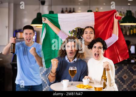 Company of emotional young adult sports fans supporting Mexican team with state flag while resting in pub with beer Stock Photo