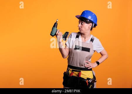 Disappointed woman contractor holding electric nail gun, posing over background. Female builder wearing overalls and hardhat in studio, using poer drilling tool for maintenance. Stock Photo