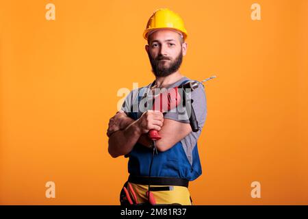 Happy building contractor holding power drill gun and posing in studio, looking at camera. Craftsman builder using drilling nail tool wearing uniform and hardhat, renovating service. Stock Photo