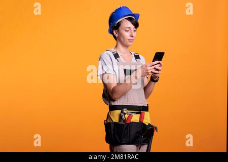 Distracted female builder using smartphone app in studio, scrolling through social media online website. Woman construction worker browsing internet network, wearing helmet and uniform. Stock Photo
