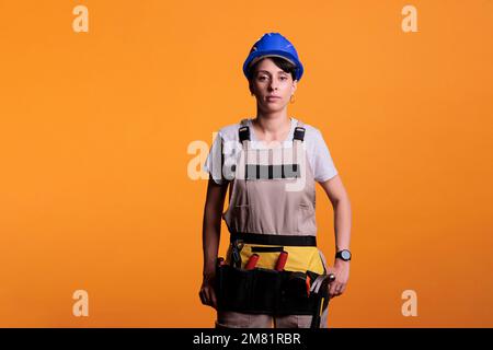 Young contractor wearing protective hardhat and looking confident at camera in studio. Confident woman construction worker posing, working in refurbishment industry over yellow background. Stock Photo
