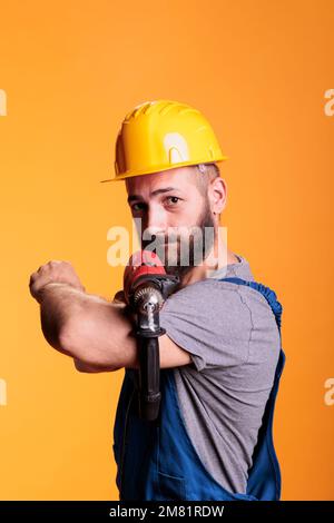 Male carpenter expert drilling holes with electric nail gun, using power drill to work on house refurbishment. Construction worker repairman holding screwing tool in studio shot. Stock Photo
