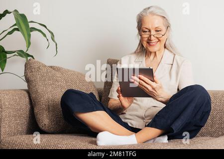 Smiling senior woman sitting with crossed legs using a digital tablet. Aged female in eyeglasses reading from a digital tablet at home. Stock Photo