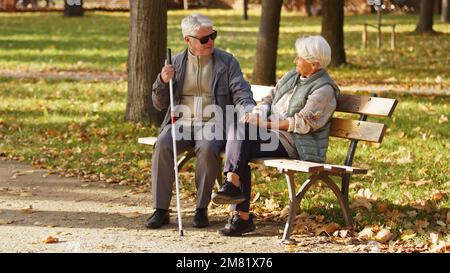 Blind senior man with sunglasses on sitting with his wife on a bench in the park. High quality photo Stock Photo
