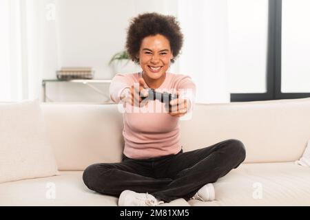 Cheerful excited young mixed race woman sitting on sofa with joystick, playing computer game in living room Stock Photo