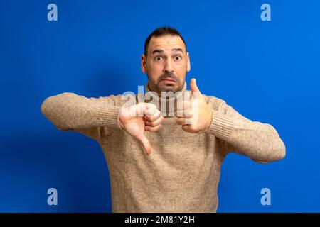 Bearded hispanic man wearing a turtleneck sweater giving a thumbs up and a thumbs down in a gesture of doubt and confusion isolated over blue Stock Photo