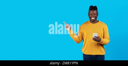 Young Black Man Holding Smartphone And Pointing Aside At Copy Space With Finger Stock Photo