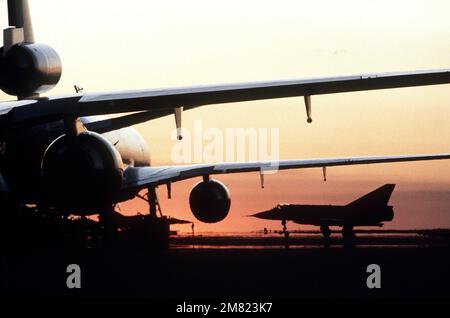 A silhouetted left side view of an RAAF Mirage III-D (French designed) aircraft parked on the flight line. Visible in the foreground is an Air Force KC-10A Extender aircraft. Both aircraft are being used during the joint Australia/New Zealand/US Exercise PITCH BLACK '84. Subject Operation/Series: PITCH BLACK '84 Base: Raaf Darwin State: Northern Territory Country: Australia (AUS) Stock Photo