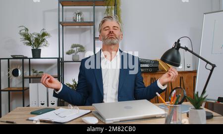 Mature businessman working on laptop computer, meditating, doing yoga breathing exercise in lotus position at home office. Calm serene freelancer old man taking break. Busy occupation. Peace of mind Stock Photo