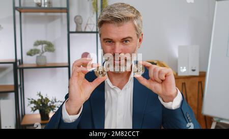 Mature businessman stock trader earning bitcoins after online monitoring trading operations. Increasing wealth, financial prosperity. Freelancer middle aged man talking to camera at office workplace Stock Photo