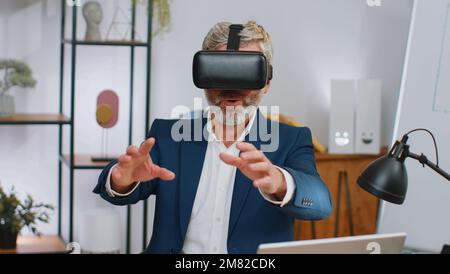 Mature businessman programmer software developer using headset helmet app to play simulation game. Watching virtual reality 3D 360 video. Mature old gray-haired in VR goggles working at home office Stock Photo