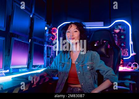 Female Asian hacker smiling in hideout place with dark environment and multiple displays and cables, trying to break into data servers and infect the system with a virus. Stock Photo