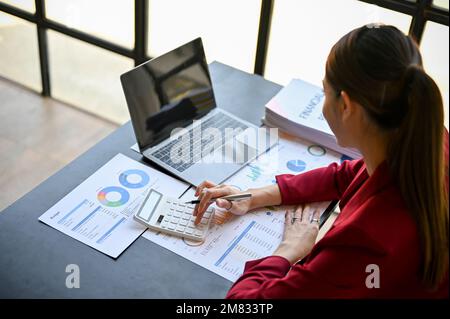 Professional millennial businesswoman or female financial analyst working on financial report, using calculator, analyzing financial data at her desk. Stock Photo