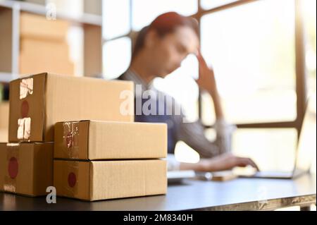 Stack of parcel boxes or cardboard boxes on the table over blurred background of a female using laptop computer. E-commerce business concept. selectiv Stock Photo