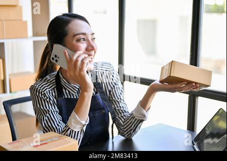 Charming and cheerful millennial Asian female online shop owner or entrepreneur talking on the phone with her suppliers while working in her packing r Stock Photo