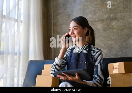 Charming millennial Asian female online seller on the phone with her supplier while working in her home living room. e-commerce business concept Stock Photo