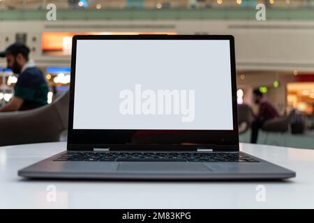 Mock up image, blank white screen laptop computer on desk in cafe or coffee shop with blurred background of people sitting inside, working from cafe. Stock Photo