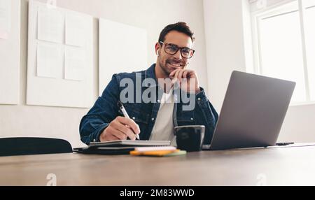 Making notes during a video conference. Happy young businessman smiling at the camera while sitting in an online meeting with a notebook. Cheerful bus Stock Photo