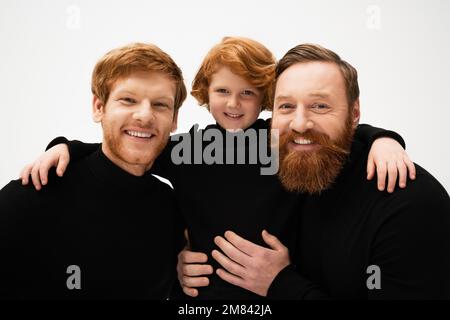 smiling redhead kid hugging bearded grandpa and dad wearing black turtlenecks isolated on grey,stock image Stock Photo